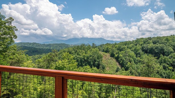 A scenic view from a wooden balcony with a metal railing, overlooking a lush green forest and distant mountains under a partly cloudy sky. - Eagle’s Nest • Epic Smoky Mtn Views • Hot Tub (Sevierville)