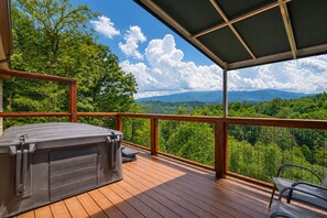 Una terraza de madera con un jacuzzi, con vistas a un paisaje boscoso y montañas a lo lejos.