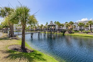 A bridge over a calm water body, surrounded by lush greenery and a row of multi-story buildings with a gazebo structure.