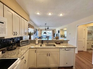 A kitchen with white cabinets, a granite countertop, a built-in dishwasher, and a window overlooking a deck.