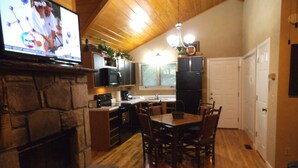 A cozy kitchen with a stone fireplace, wooden dining table, and a TV mounted on the wall. - Dove's Nest: Beautiful, Pet Friendly Cabin Alongside a Creek near Downtown Gatlinburg (Gatlinburg)
