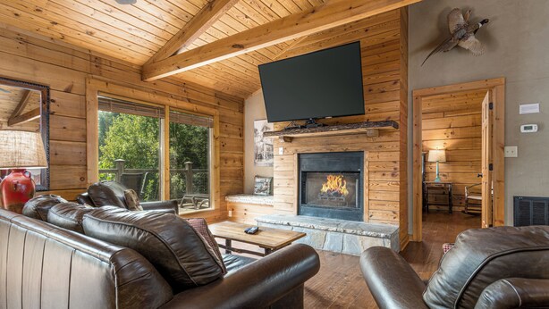 A log cabin interior with a fireplace, leather sofas, and a television mounted on the wall.