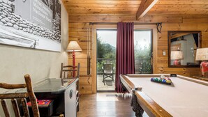 A wooden interior with a pool table, a table with a red lamp, and a view of the outdoors through a glass door.