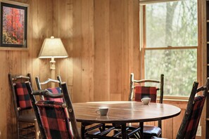 Un intérieur en bois avec une table ronde, des chaises recouvertes d’un tissu à carreaux, une lampe et une fenêtre donnant sur des arbres.