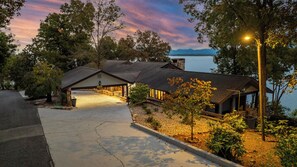A lakeside cabin with a driveway leading to a covered porch, surrounded by trees and a view of the lake.