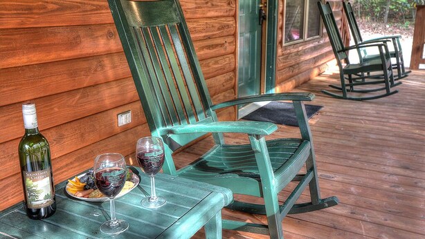 Una terraza de madera con una mesa, dos mecedoras y una botella de vino.