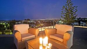 Een balkon met rieten meubilair, twee kaarsen en een uitzicht op een stadslandschap in de schemering.