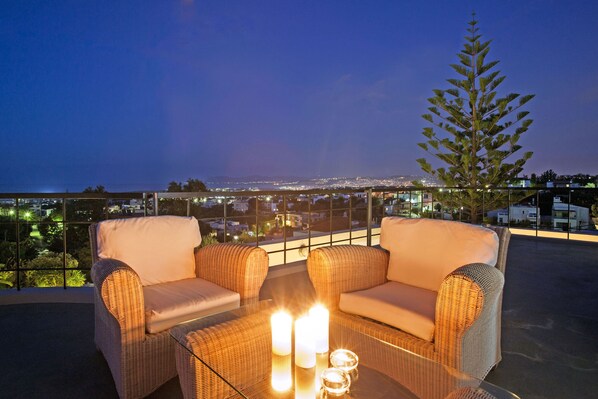 A balcony with wicker furniture, two candles, and a view of a cityscape at dusk. - Villa Zaimis - Five Bedroom Villa, Sleeps 10 (Daratsos)