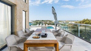 Un balcón con mobiliario de mimbre, una mesa con platos y vasos, y una vista a una piscina y edificios a lo lejos.
