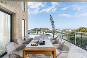 A balcony with wicker furniture, a table with plates and glasses, and a view of a pool and buildings in the distance.
