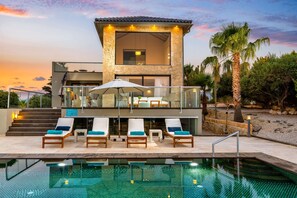 A modern two-story building with a pool area, outdoor seating, and a view of the sunset.