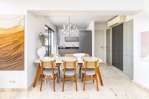 A modern dining area with a white table and wicker chairs, a kitchen in the background, and a large artwork on the left wall.