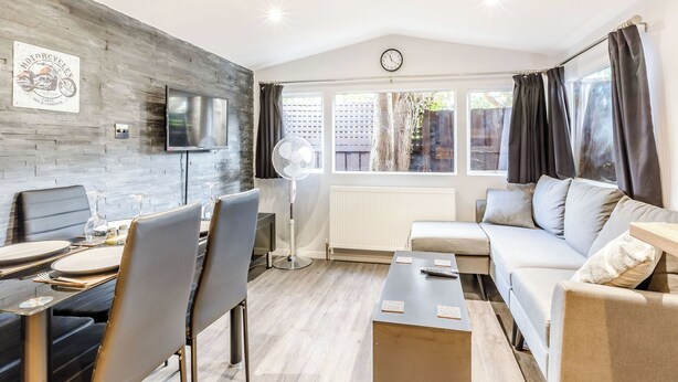 A modern living room with a dining area, a sofa, a TV mounted on a brick wall, and a window with curtains. - 2 bedroom accommodation in Abbotskerswell, nr Newton Abbot (Abbotskerswell, nr Newton Abbot)