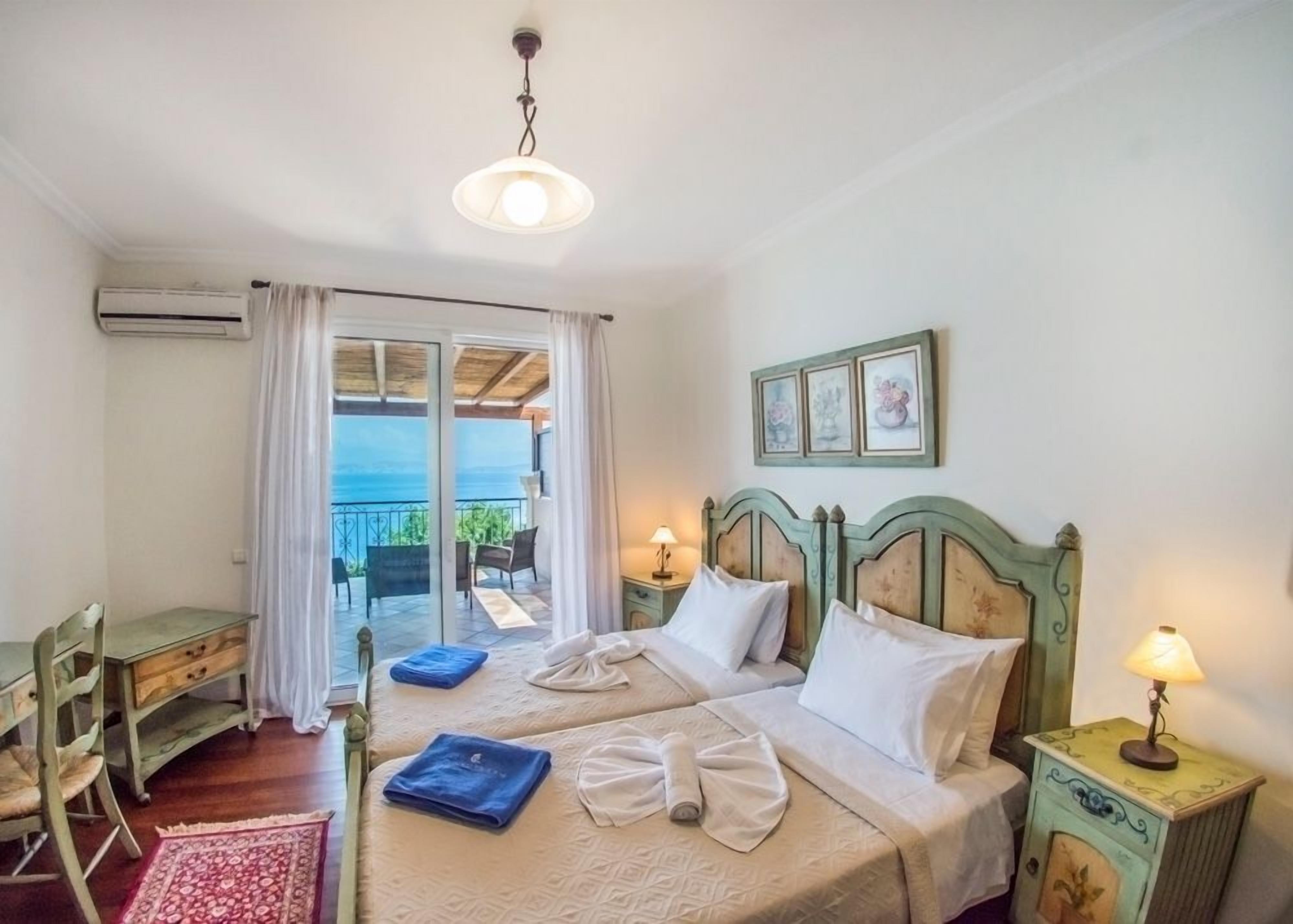 A bedroom with a bed, a desk, a chair, a lamp, and a view of the sea through a glass door.