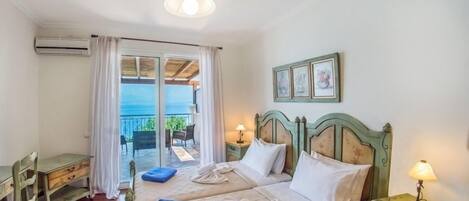 A bedroom with a bed, a desk, a chair, a lamp, and a view of the sea through a glass door.