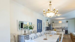 A dining area with a long table, chairs, and a chandelier. There is a kitchen in the background with cabinets and a refrigerator. - Villa Aliki - Five Bedroom Villa, Sleeps 10 (Kassiopi)