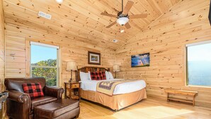 A wooden cabin room with a bed, a leather sofa, a ceiling fan, and large windows.
