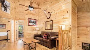 A wooden cabin interior with a brown leather sofa, a wooden coffee table, a ceiling fan, and a mounted deer head.
