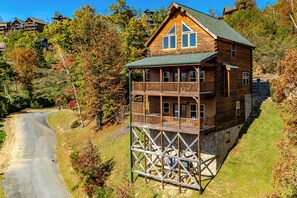 En træhytte med balkon på en skråning, omgivet af træer og en vej.