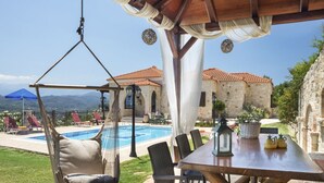 A poolside area with a hanging chair, a table with chairs, and a view of a stone building with a tiled roof.