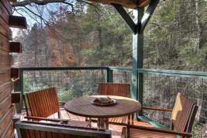 Une terrasse en bois avec une table ronde et des chaises, entourée d’une rambarde verte, donnant sur une zone boisée.