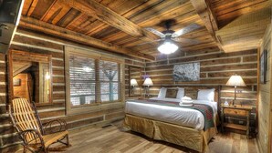 Quarto de cabana de madeira com uma cama, duas luminárias de apoio, uma cadeira de balanço e um ventilador de teto.