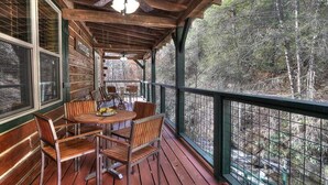 Um deck de madeira com mesa e cadeiras, um ventilador de teto e vista para uma área arborizada.