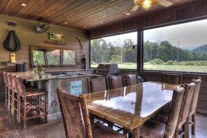 A rustic bar area with a wooden table and bar stools, a grill, and a view of the outdoors.