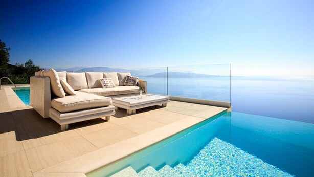 Una piscina infinita moderna con vista despejada al mar, un sofá color beige con cojines y una mesita con un florero.