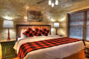 A wooden cabin-style bedroom with a bed, two bedside lamps, a painting on the wall, and a window with blinds. - Walk to Downtown-NEW Game Room-Hot Tub-2 Kings (Gatlinburg)