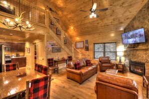 Interior espacioso de una cabaña con comedor, chimenea, televisor y una escalera que lleva a un nivel superior.