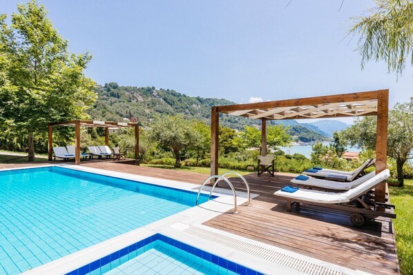 A swimming pool with lounge chairs and a pergola, surrounded by greenery and a scenic view.