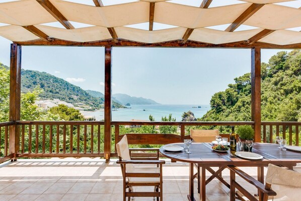 A wooden deck with a table set for dining, overlooking a scenic coastal landscape with hills and the sea.