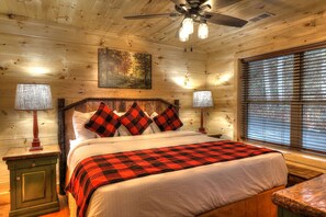A cozy wooden cabin bedroom with a bed, two bedside lamps, a ceiling fan, and a window with blinds.