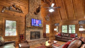 A log cabin interior with a stone fireplace, leather furniture, and mounted animal heads.