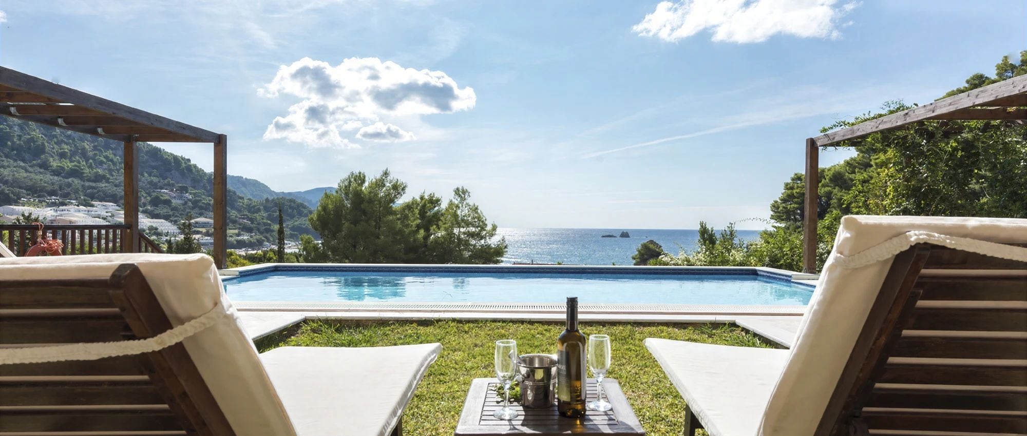 A poolside view with lounge chairs, a table with glasses and a bottle, and a view of the sea.