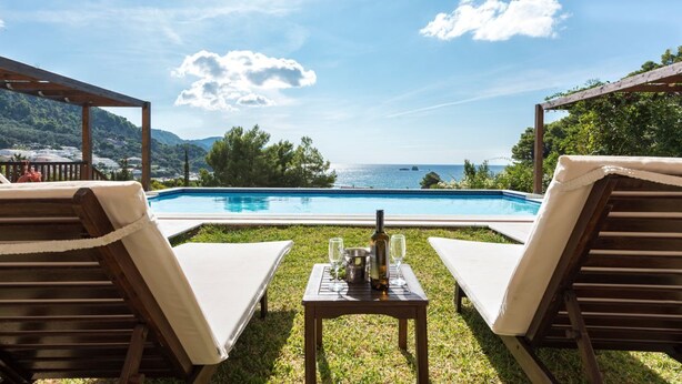 A poolside area with two lounge chairs, a table with a bottle and glasses, and a view of the sea and mountains.