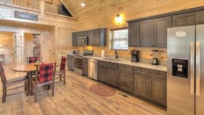 Interior de una cabaña de madera con una zona de cocina que incluye armarios oscuros, electrodomésticos de acero inoxidable y una mesa de comedor con sillas a cuadros rojos y negros.