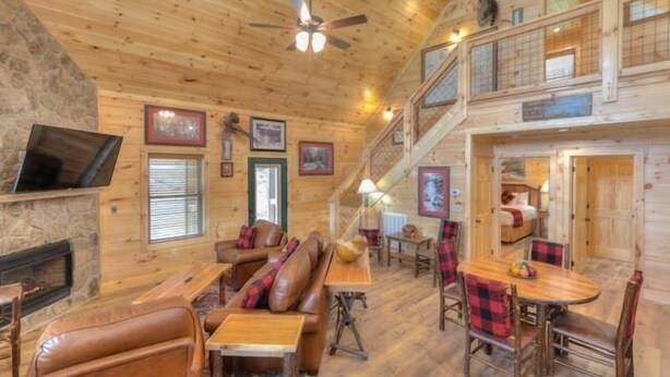 Interior de una cabaña de madera con chimenea, sofá de cuero, mesa de comedor y una escalera a un nivel superior.