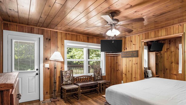 Ein Zimmer mit Holzwänden, ausgestattet mit einem Bett, einem Schreibtisch, einem Stuhl, einem Deckenventilator und einem an der Wand befestigten Fernseher.