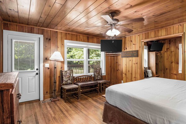 A wooden-paneled room with a bed, a desk, a chair, a ceiling fan, and a TV mounted on the wall.