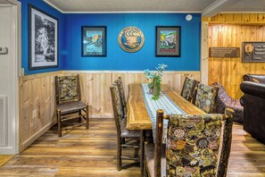 A dining area with a wooden table, patterned chairs, and a blue wall with framed pictures and signs. - Poppy’s Outpost • Hot Tub • Fire Pit • Family Fun (Cosby)