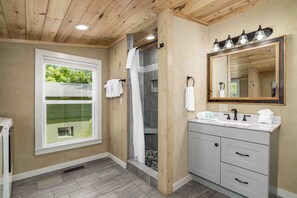 A bathroom with a shower, a vanity with a mirror, and a window with a view of greenery.