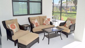 Un patio iluminado por el sol con muebles de mimbre, incluyendo un sofá y sillones, junto con una mesa de centro de vidrio.