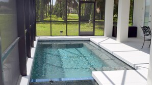 Piscina con cerramiento de vidrio, rodeada de muros blancos y vista a un área verde.