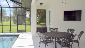 Zona junto a la piscina con una mesa redonda y sillas, un televisor montado en la pared, y vistas al exterior.