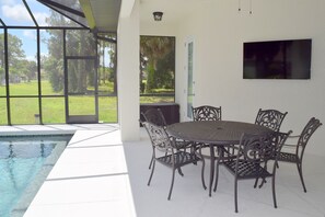 A poolside area with a round table and chairs, a TV mounted on the wall, and a view of the outdoors. - NEW! 3/3/2 HEATED POOL HOME IN ROTONDA WEST (Rotonda West)