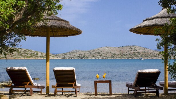 Eine Strandlounge mit Holzliegen und Strohdächern, mit Blick auf ein ruhiges Meer und sanfte Hügel in der Ferne.