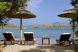 A beachside lounge with wooden lounge chairs and thatched umbrellas, overlooking a calm sea and distant hills.