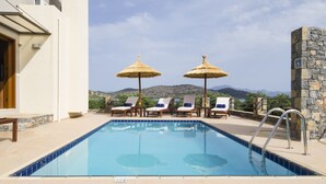Ein Schwimmbad mit Liegestühlen und Sonnenschirmen, eine Steinmauer und Blick auf Hügel.